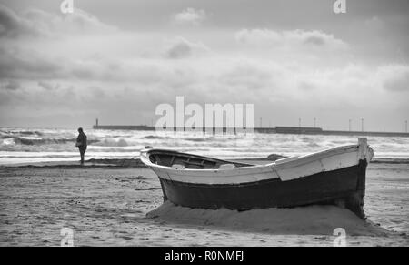 Moody black and white photo of an abandoned boat stuck in the sand on a beach with a single person in the distance near the shore with waves and cloud Stock Photo