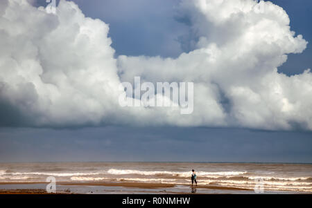 Unidentifiable single woman walking on a brown sand beach with waves crashing and ominous clouds overhead in a grey and blue sky Stock Photo