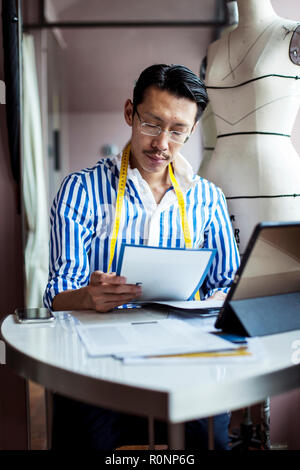 Japanese male fashion designer working in his studio, sitting at table, looking at digital tablet. Stock Photo