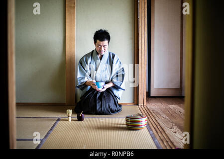 Japanese man wearing traditional kimono kneeling on tatami mat, holding tea bowl, during tea ceremony. Stock Photo