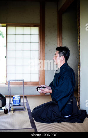 Japanese man wearing traditional kimono kneeling on floor, holding tea bowl, during tea ceremony. Stock Photo