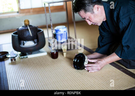 Japanese man wearing traditional kimono kneeling on floor, holding tea bowl, during tea ceremony. Stock Photo