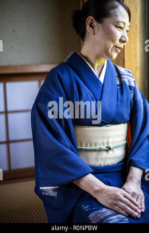 Japanese woman wearing traditional bright blue kimono with cream coloured obi kneeling on floor in traditional Japanese house.