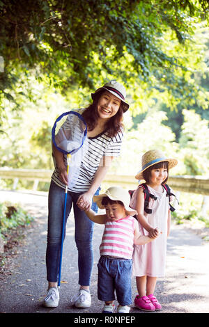 Smiling Japanese woman holding butterfly net and two girls wearing sun hats standing on path, looking at camera. Stock Photo