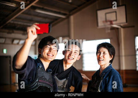 Smiling female and two male Japanese Kendo fighters standing in a gym, taking selfie with mobile phone. Stock Photo