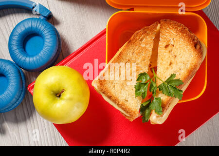 Yellow sandwich box with toasted slices of bread, cheese and green parsley, green apple, headphones and hardback books on the background. School break Stock Photo