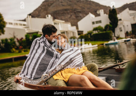 Man kissing his girlfriend sitting in a boat while on a date at a lake. Young couple wrapped in a towel sitting in a boat relaxed spending time togeth Stock Photo