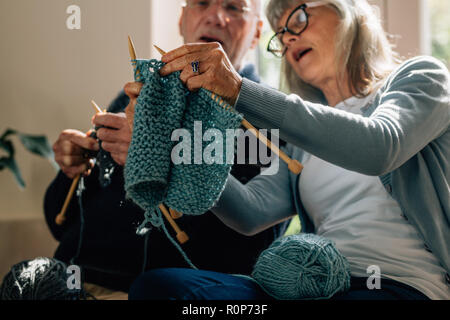 Senior woman teaching her husband the art of knitting woollen clothes. Senior man learning to knit woollen clothes from his wife sitting at home. Stock Photo