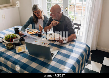 Senior man and woman having breakfast together at home with a laptop and papers on the table. Woman holding a paper discussing finances with her husba Stock Photo