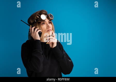 Steam punk girl wearing glasses uses a walkie talkie and poses as thinking. Dark hoodie with long sleeves shot against blue background Stock Photo