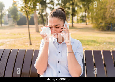 Sad young woman cries while talking on the mobile phone in the park on bench Stock Photo