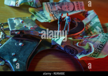 Conceptual miniature criminal businessmen shake hands standing on top of handcuffs with crumpled dollar bills and police lights Stock Photo