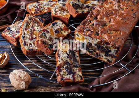delicious chunky dried fruits rich cake on a wire cake stand with brown cloth, cinnamon sticks, dried apricots and date fruits on a rustic wooden tabl Stock Photo