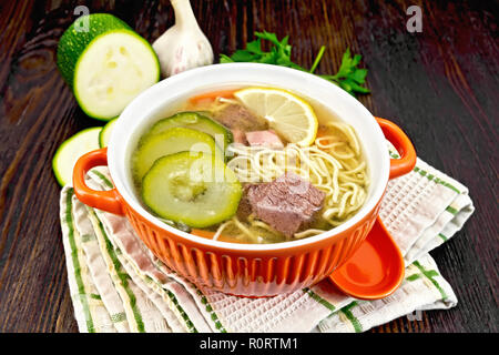 Soup with zucchini, beef, ham, lemon and noodles in a bowl, parsley and dill on a towel on a wooden board background Stock Photo