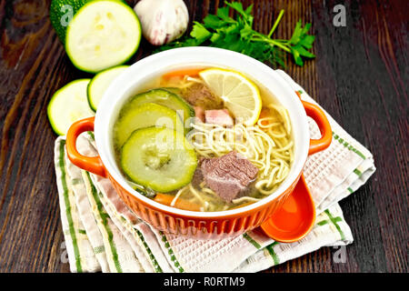 Soup with zucchini, beef, ham, lemon and noodles in a bowl, parsley and dill on a towel against a dark wooden board Stock Photo