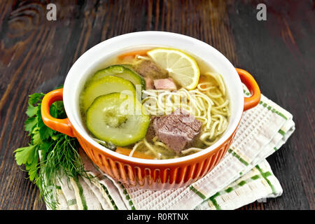 Soup with zucchini, beef, ham, lemon and noodles in a red bowl, parsley and dill on a napkin on a wooden board background Stock Photo