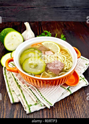 Soup with zucchini, beef, ham, lemon and noodles in a bowl, parsley and dill on a napkin on a wooden board background Stock Photo