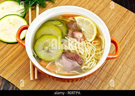 Soup with zucchini, beef, ham, lemon and noodles in a bowl, parsley and dill on a bamboo napkin against a wooden board Stock Photo