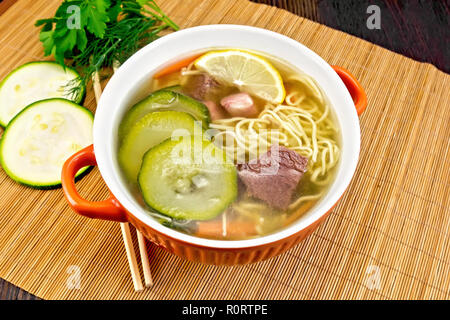 Soup with zucchini, beef, ham, lemon and noodles in a bowl, parsley and dill on a bamboo napkin against a dark wooden board Stock Photo