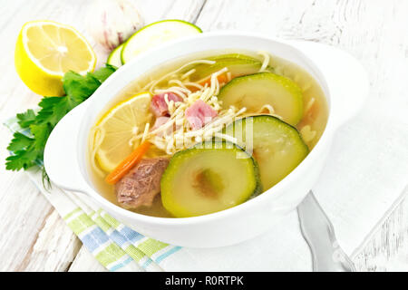 Soup with zucchini, beef, ham, lemon and noodles in a bowl on a towel, parsley and dill on a wooden plank background Stock Photo