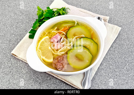 Soup with zucchini, beef, ham, lemon and noodles in a bowl on a napkin, parsley and dill on a gray granite table background Stock Photo