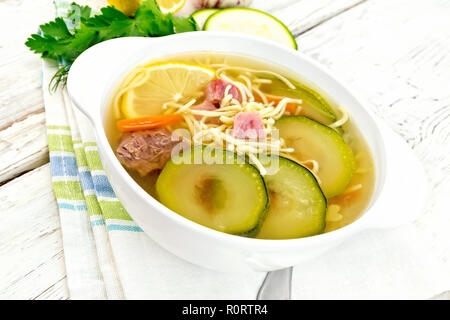 Soup with zucchini, beef, ham, lemon and noodles in a bowl on a napkin, parsley and dill on a light wooden board background Stock Photo