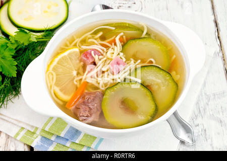 Soup with zucchini, beef, ham, lemon and noodles in a bowl on a napkin, parsley and dill on a wooden plank background Stock Photo