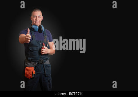 Strong and confident male mechanic in blue overal standing with orange gloves, white helmet, ear protection and tools on a black background Stock Photo