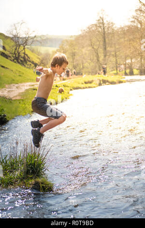 A little boy with ADHD, Autism, Aspergers Syndrome jumps into the river Dove at Dovedale on a hot summers day in denim shorts and hiking boots