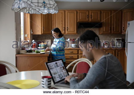 Latinx man working at laptop with senior mother cooking in kitchen
