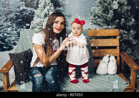 Portrait of loving mother decorating Christmas tree with her toddler girl sitting beside gifts on the floor while snow falling down. Smiling at each o Stock Photo