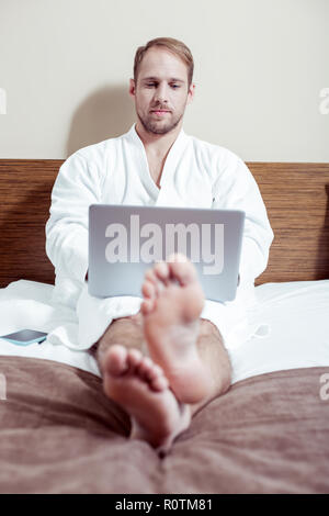 Bearded pleasant man holding his silver laptop working in bed Stock Photo