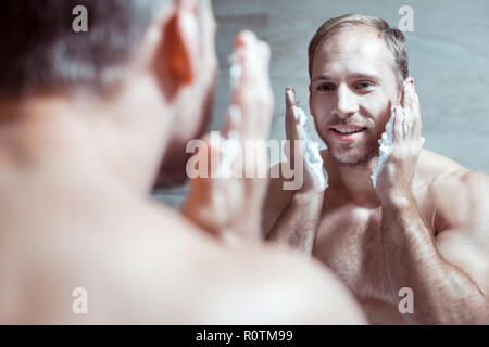 Cheerful blue-eyed man smiling while looking into mirror and shaving face Stock Photo