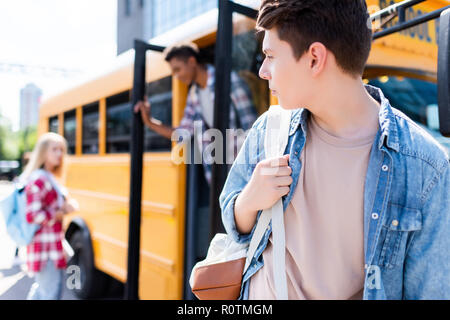 teen schoolboy walking in front of school bus and turning back at classmates Stock Photo