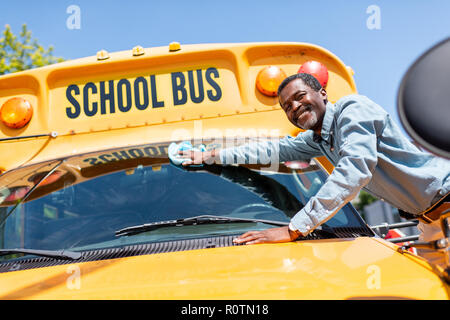 smiling mature african american bus driver wiping front window of school bus and looking at camera