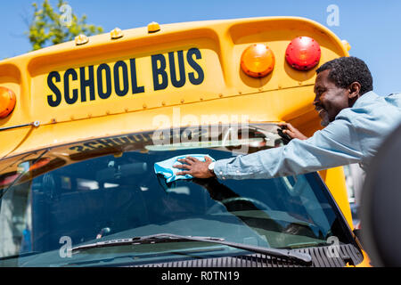 happy mature african american bus driver wiping front window of school bus