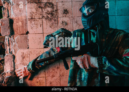 male paintball player in camouflage and goggle mask hiding behind wall and holding marker gun in abandoned building Stock Photo