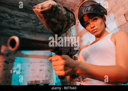 attractive female paintballer in camouflage and goggle mask above head aiming by paintball gun in abandoned building Stock Photo
