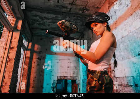 low angle view of young female paintballer in camouflage and white singlet aiming by marker gun in abandoned building Stock Photo