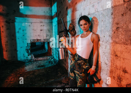 confident young female paintballer in camouflage and white singlet holding marker gun in abandoned building Stock Photo