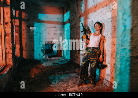 young female paintballer in camouflage and white singlet holding paintball gun and goggle mask in abandoned building Stock Photo