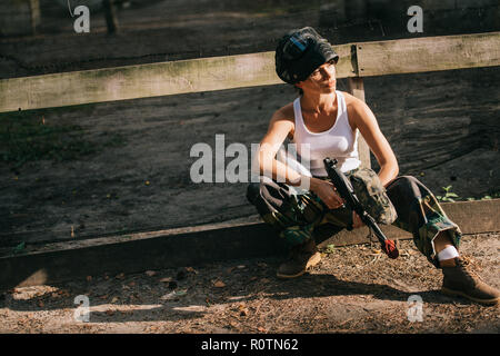 confident young female paintballer in white singlet and goggle mask holding paintball gun outdoors Stock Photo