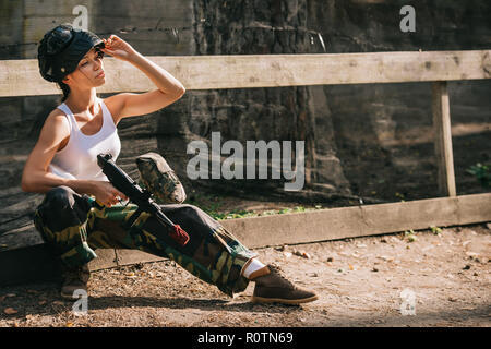 young female paintballer in white singlet putting on goggle mask and holding paintball gun outdoors Stock Photo