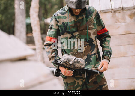 male paintball player with marker gun looking at camouflage covered by paintball splash outdoors Stock Photo