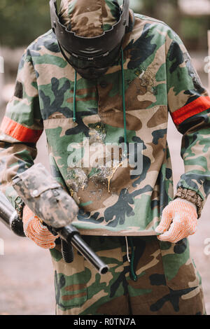 paintball player with marker gun looking at camouflage covered by paintball splash outdoors Stock Photo