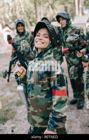 attractive smiling female paintballer in uniform holding paintball gun while her team standing behind outdoors Stock Photo