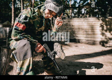 smiling young female paintballer in camouflage uniform sitting with paintball gun outdoors Stock Photo