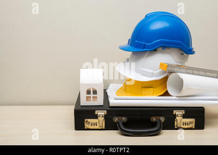 Engineering accessories, helmets, house paper model, business bag, blueprint, on wood table with white concrete wall background. Picture for add text  Stock Photo