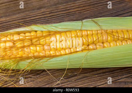 Close-up of corn cob in green husk. Zea mays. Beautiful ripe corncob detail. Golden maize grains, silks, thin leaves. Natural brown wooden background. Stock Photo