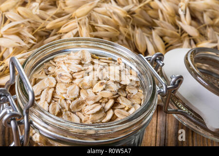 Glass jar full of oat flakes. Common oats. Avena sativa. Organic rolled oats. Healthy nutrients, dietary fiber. Brown wooden board. Dry cereal grains. Stock Photo